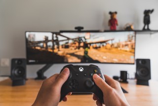 A person holding an xbox controller in the foreground with a blurred monitor in the background.